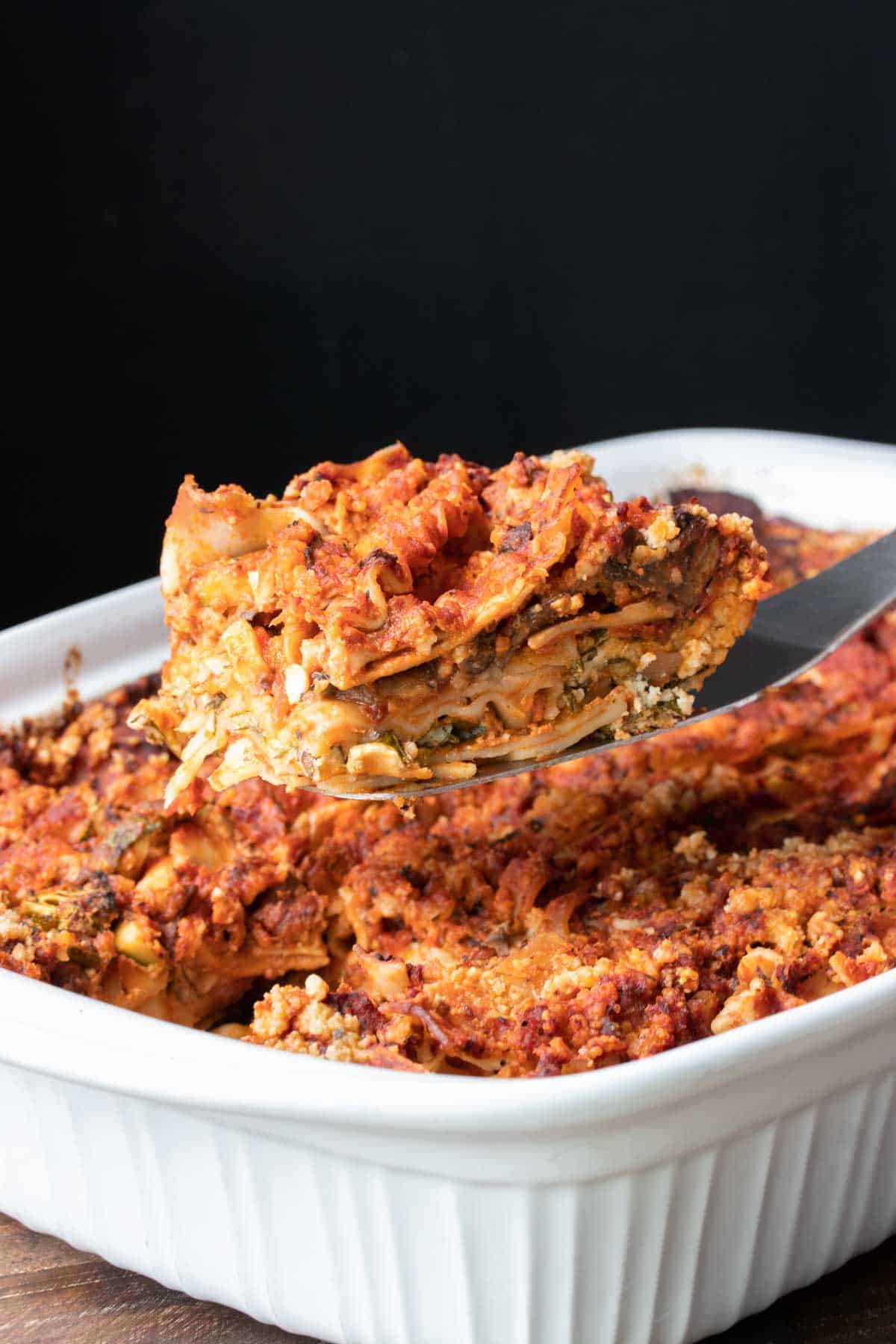 zilveren spatel die een stuk plantaardige lasagne uit een witte ovenschaal haalt