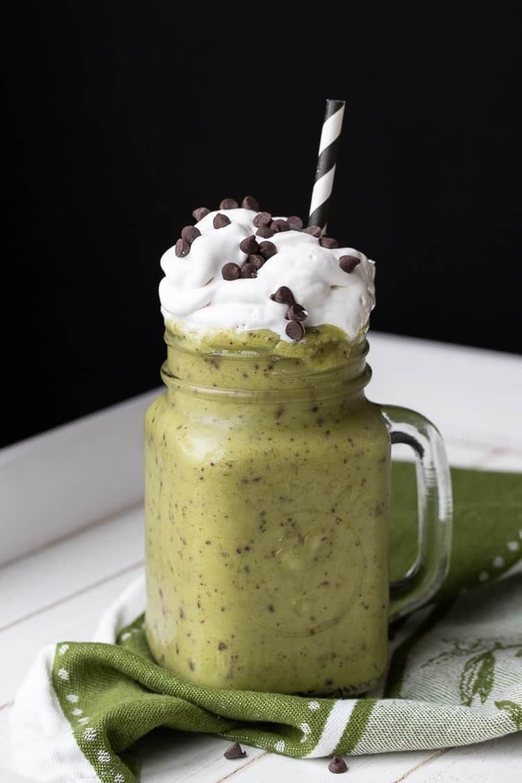 Green shake with chocolate chips in glass jar topped with whipped cream and a straw