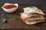 A stack of lasagna wontons on a wooden surface next to dipping sauce