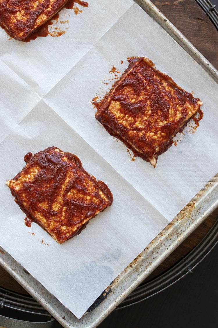 Squares of tempeh slathered in BBQ sauce on a parchment lined cookie sheet