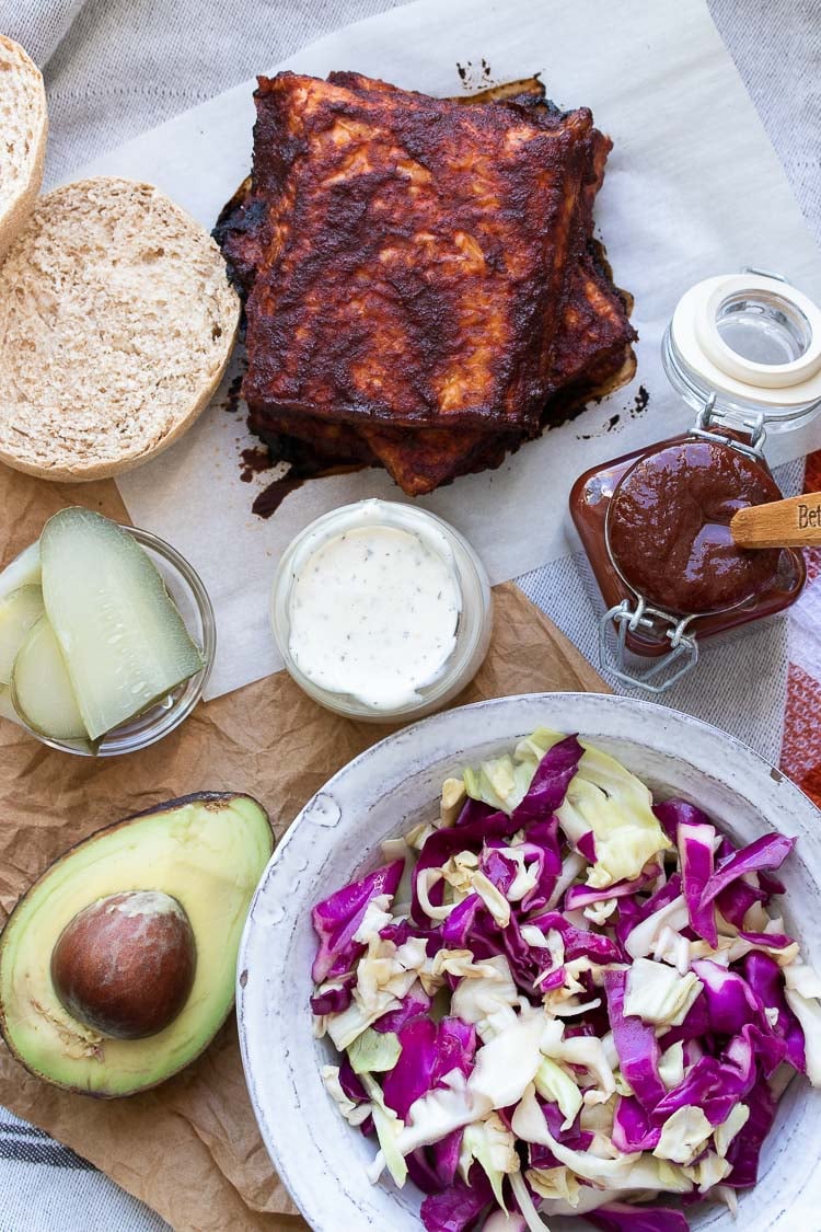 Bread roll, BBQ tempeh squares, shredded cabbage, avocado, pickles, ranch and BBQ sauce on a table