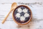 Top view of a smoothie bowl in a wooden bowl with a design on top that looks like a soccer ball made of blueberries and coconut.