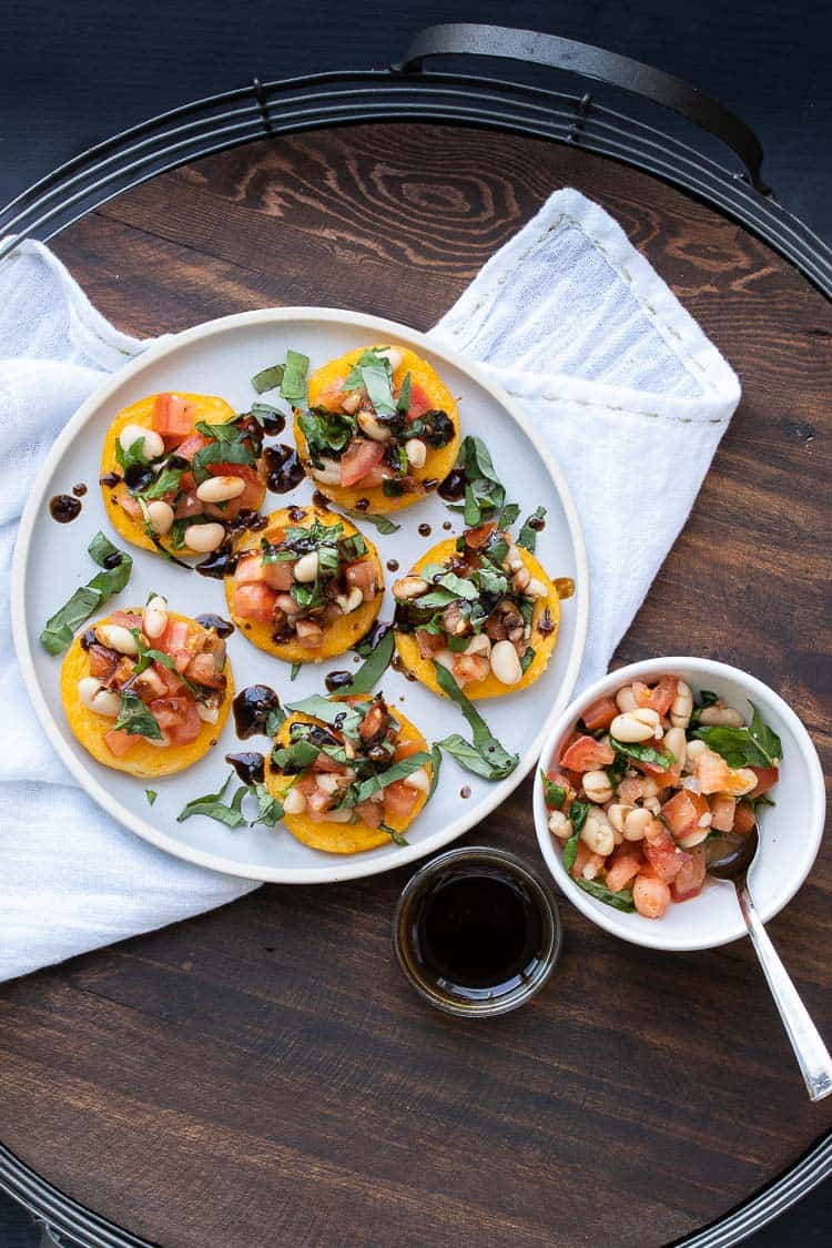 Bruschetta topped polenta rounds on a round white plate set on a wooden tray