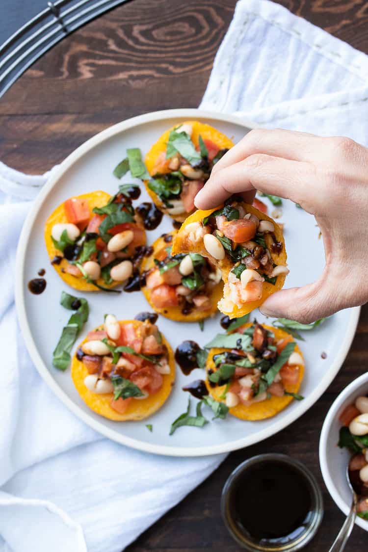 Hand picking up a half eaten bruschetta topped polenta round from a white plate