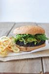 A black rice burger on a wooden surface with onion rings at the side