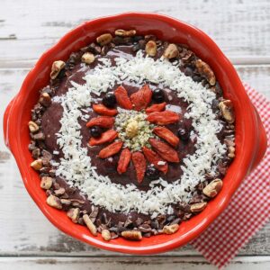 Top view of a smoothie bowl in a red bowl topped with goji berries, nuts, coconut and hemp seeds in a geometric design.
