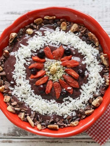 Top view of a smoothie bowl in a red bowl topped with goji berries, nuts, coconut and hemp seeds in a geometric design.