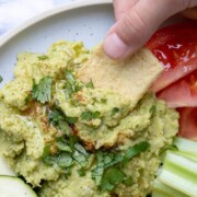 Hand dipping pita chip into a pile of hummus topped with oil and parsley