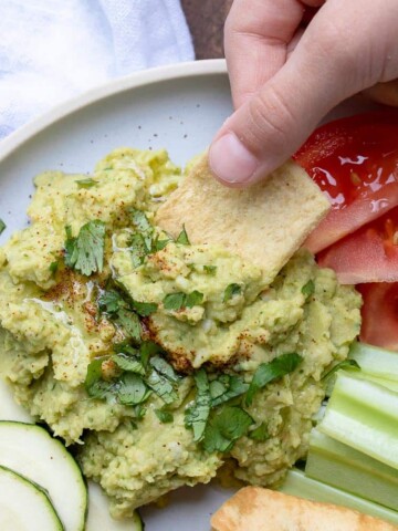 Hand dipping pita chip into a pile of hummus topped with oil and parsley