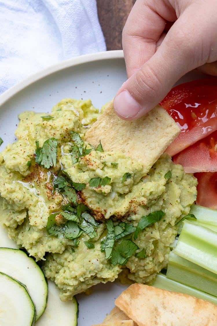 Hand dipping pita chip into a pile of hummus topped with oil and parsley