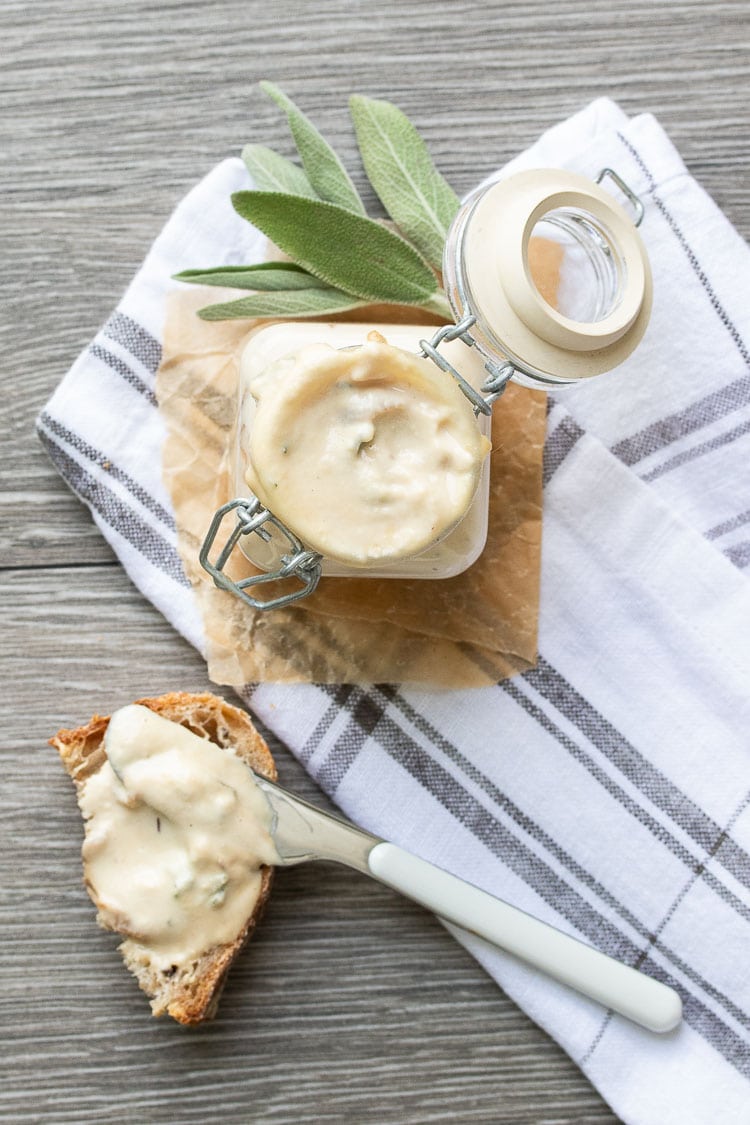 Glass jar filled with tan creamy sauce next to knife spreading sauce on bread piece
