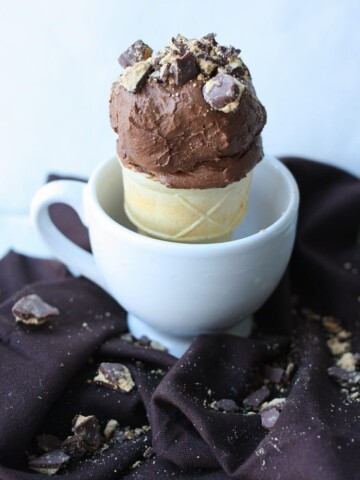 Chocolate ice cream topped with peanut butter cup crumbles in a cone sitting in a white mug.