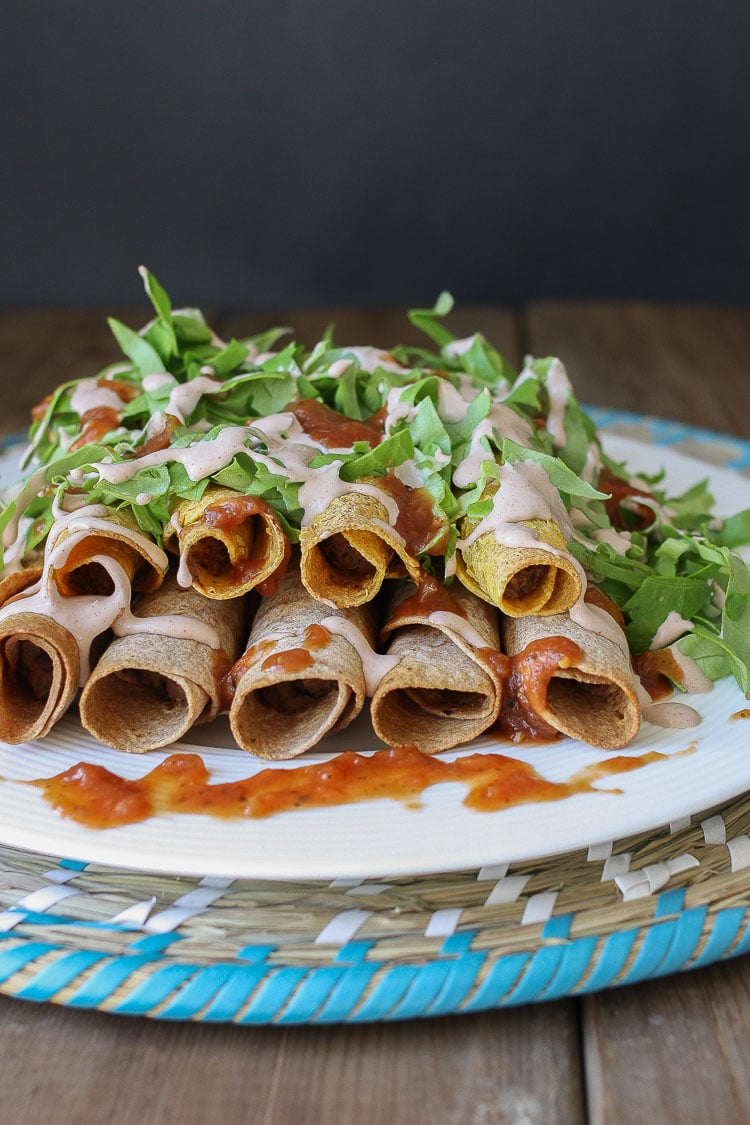 A photo of Rolled Tacos on a plate with arugula on top and drizzled with hot sauce