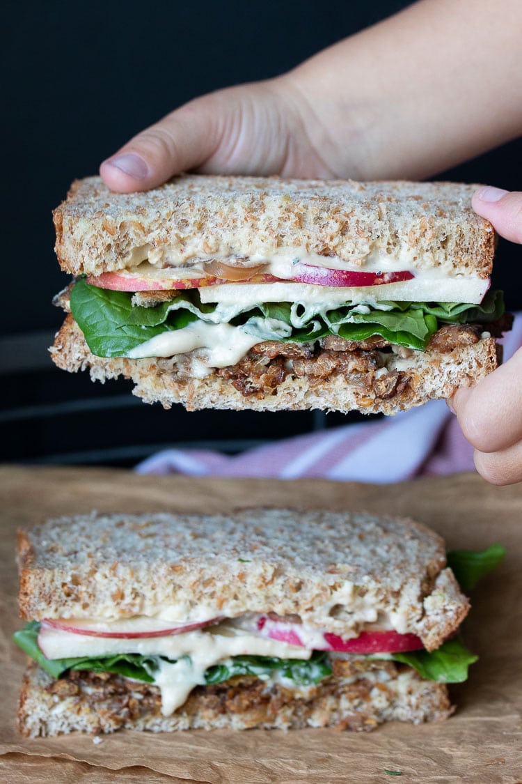 Hands taking half a sandwich with tempeh bacon, arugula and apples off of the other half