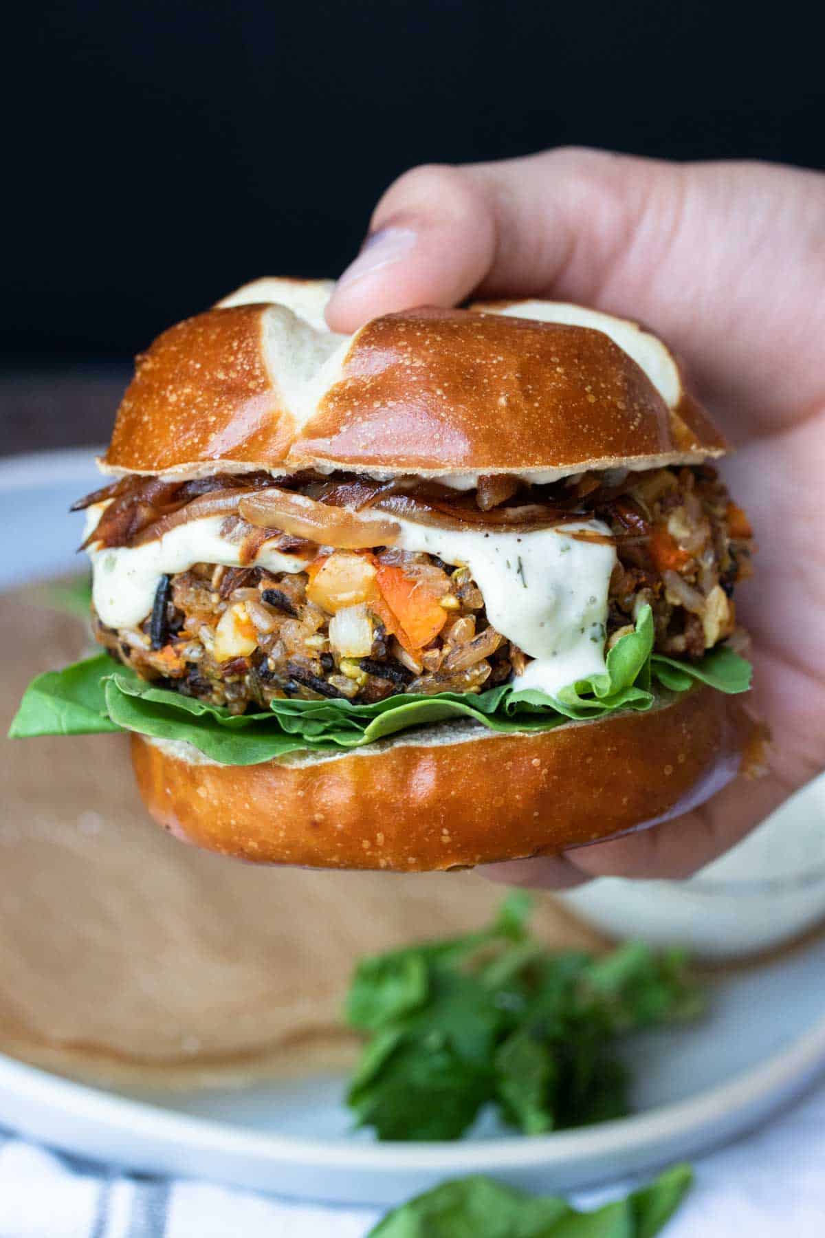 Hand holding a butternut squash burger on a pretzel bun with onions, lettuce and ranch.