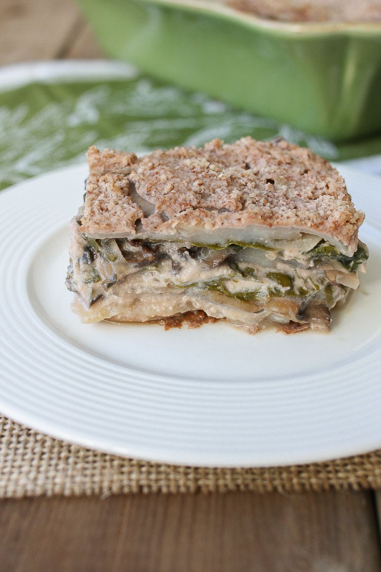 A slice of veggie casserole sitting on a white plate white a green baking dish in the background