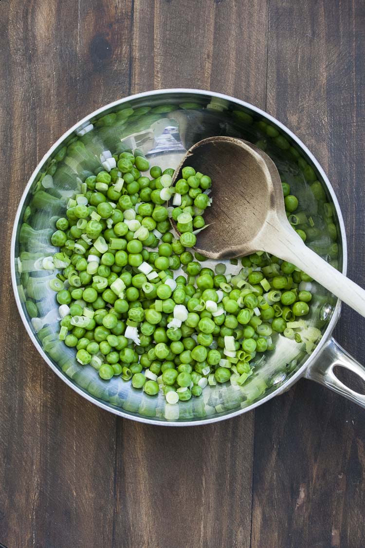 Peas and chopped scallions being mixed in a metal pan with a wooden spoon