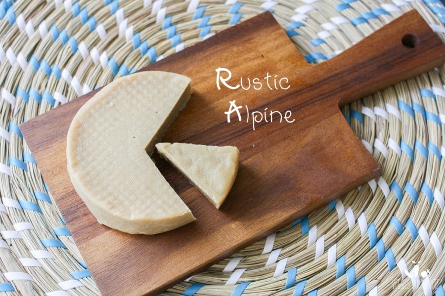 A photo of rustic alpine Miyoko's Creamery Vegan Cheese sitting on a wooden chopping board with a slice cut out