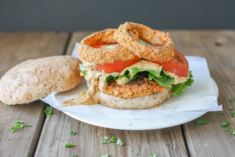 A side shot of a chickpea burger topped with lettuce and tomato sitting on a white plate with herbs scattered at the side