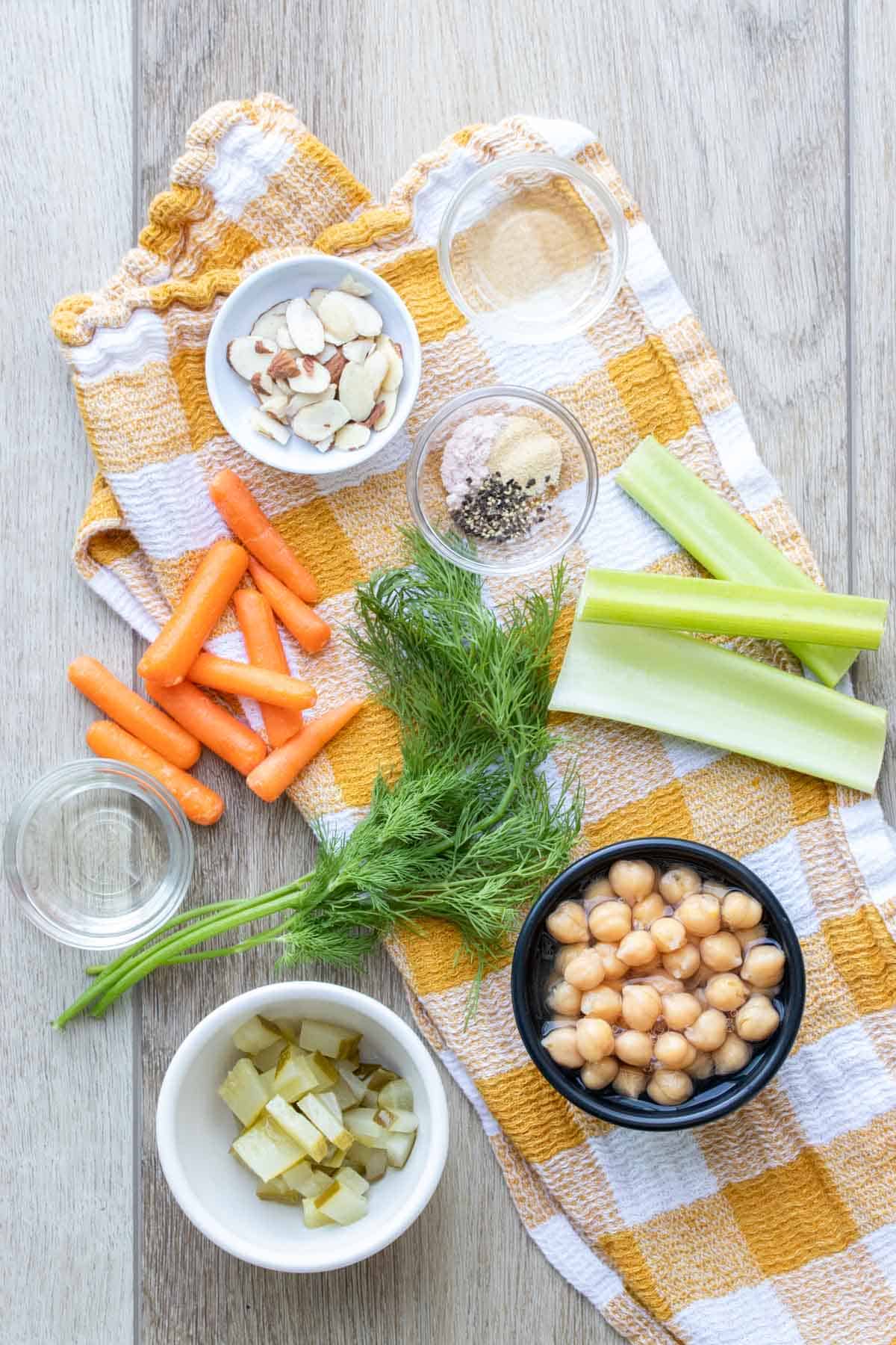 Ingredients needed to make a mashed chickpea salad sandwich on a checkered towel on a wooden surface