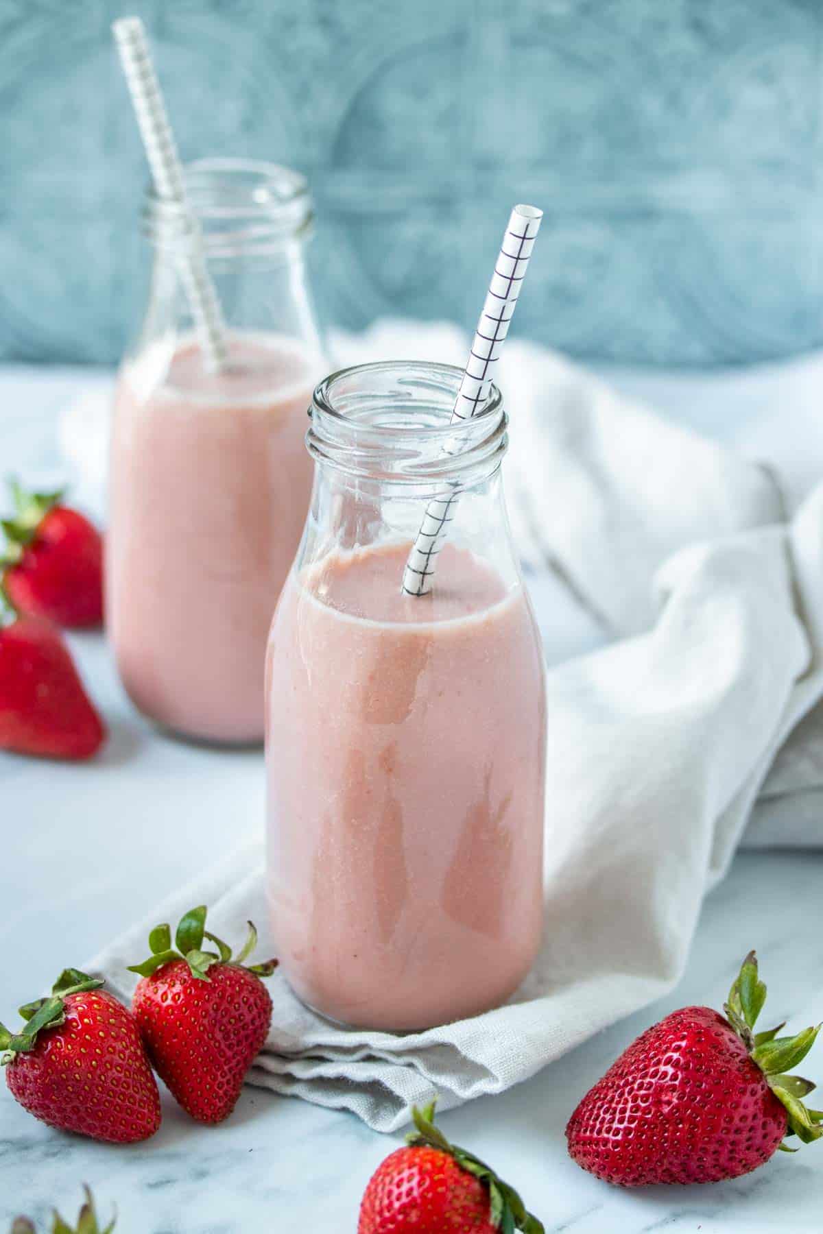 An angled top view of two glass jars with a pink milk in them on a light towel surrounded by strawberries