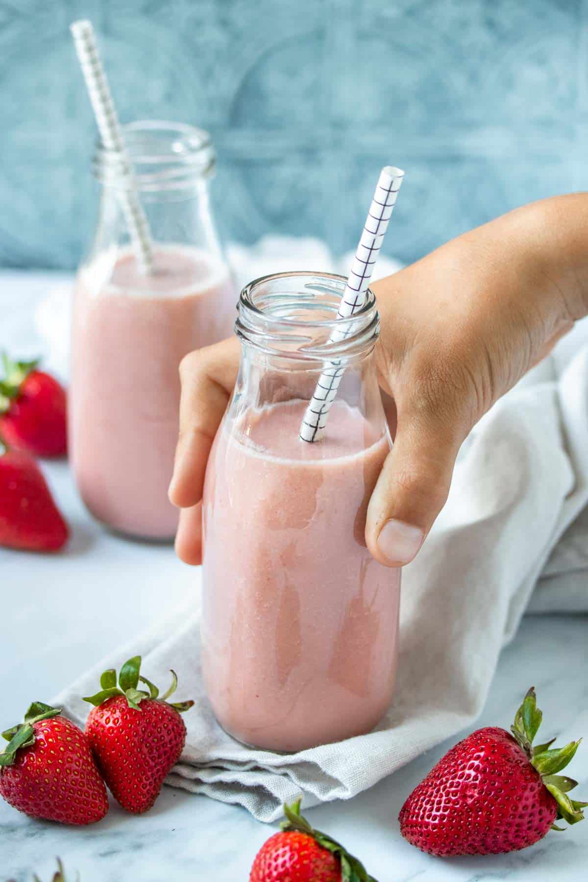 A hand holding a milk jar filled with a pink milk on a light colored towel with strawberries on it