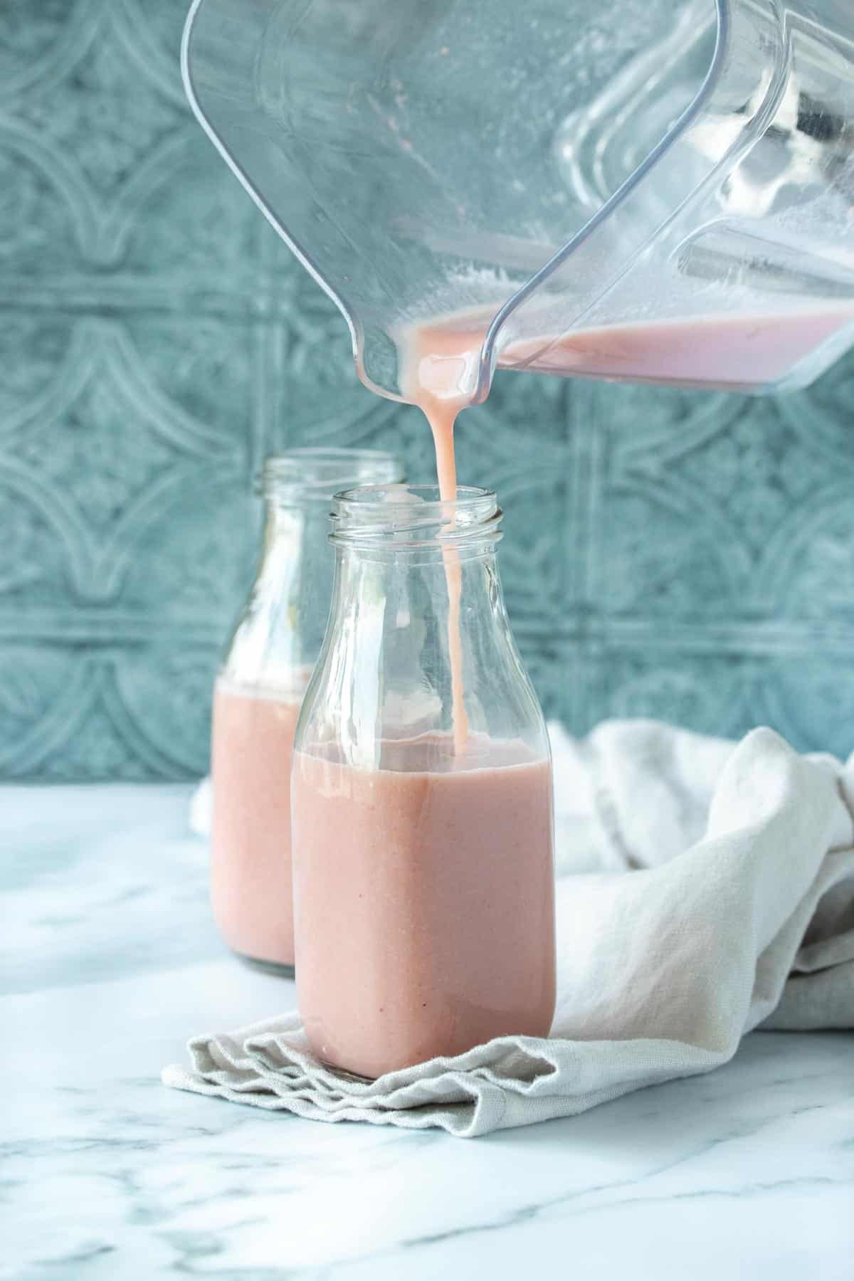 A blender pouring a pink milk into a glass milk jar in front of a full jar