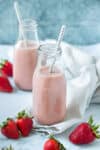 Two glass milk jars on a white surface filled with a pink milk surrounded by strawberries