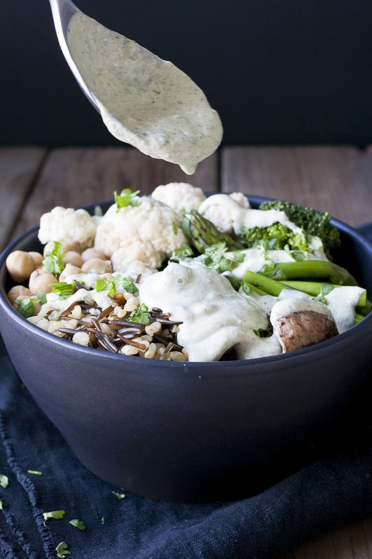 Spoon drizzling white sauce on top of veggies, rice and beans in a black bowl