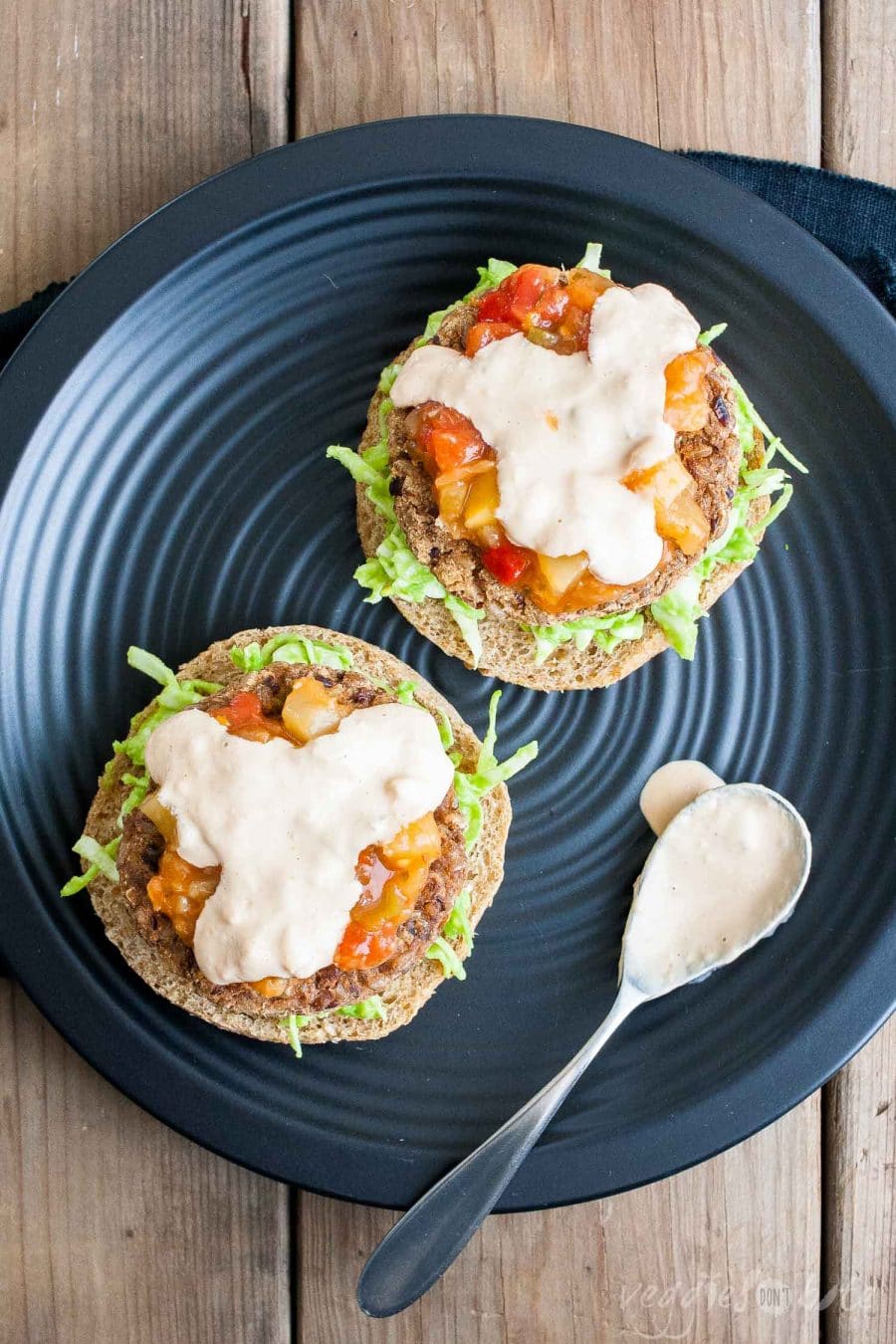 AN overhead shot of open vegan black bean burgers drizzled with sauce with a small spoon at the side