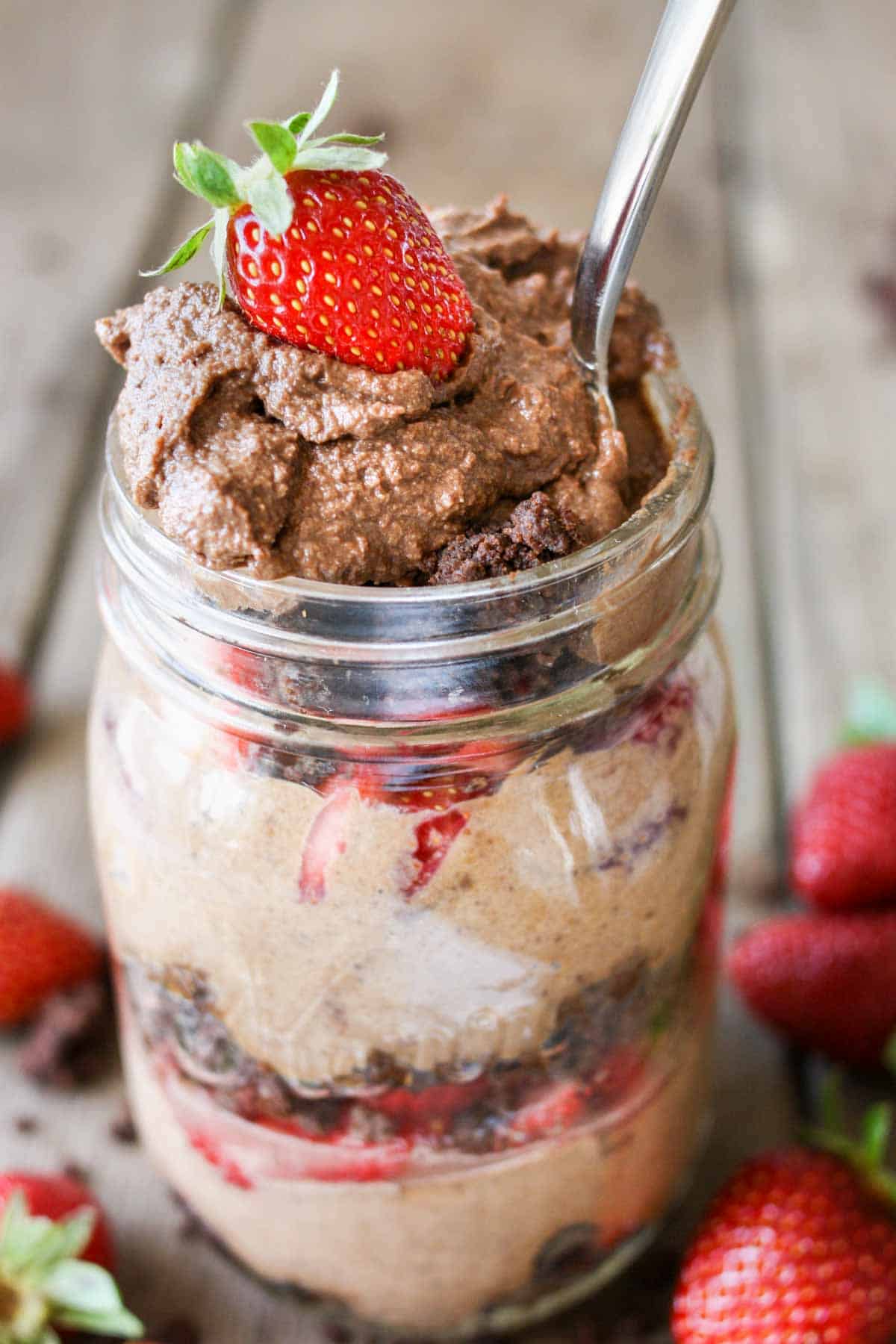 A spoon in a glass jar filled with chocolate mousse, strawberries and brownie crumbles.
