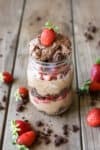 A glass mason jar that has chocolate mousse in it layered with brownies crumbled up and strawberries sitting on a wooden surface.