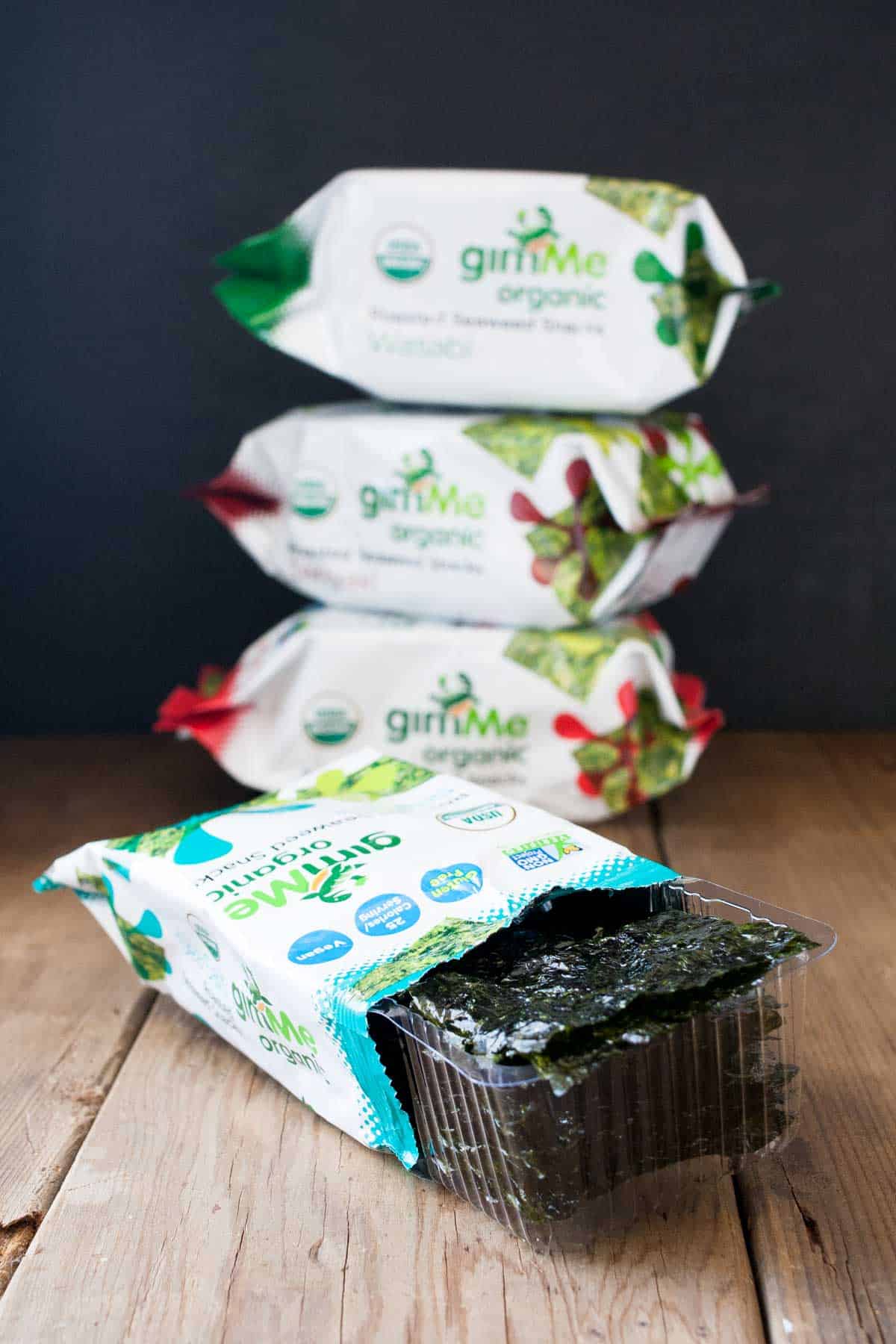 An open seaweed snack packet with the snacks coming out in front of a pile of three of them.