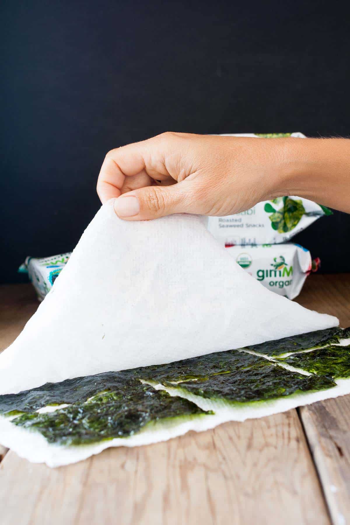 A hand lifting a paper towel over a layer of seaweed snacks lined up on another paper towel.