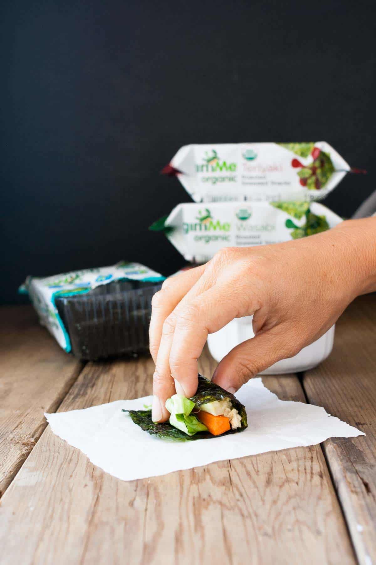 A hand rolling a seaweed snack filled with veggies and rice on top of a paper towel sitting on a wooden surface.