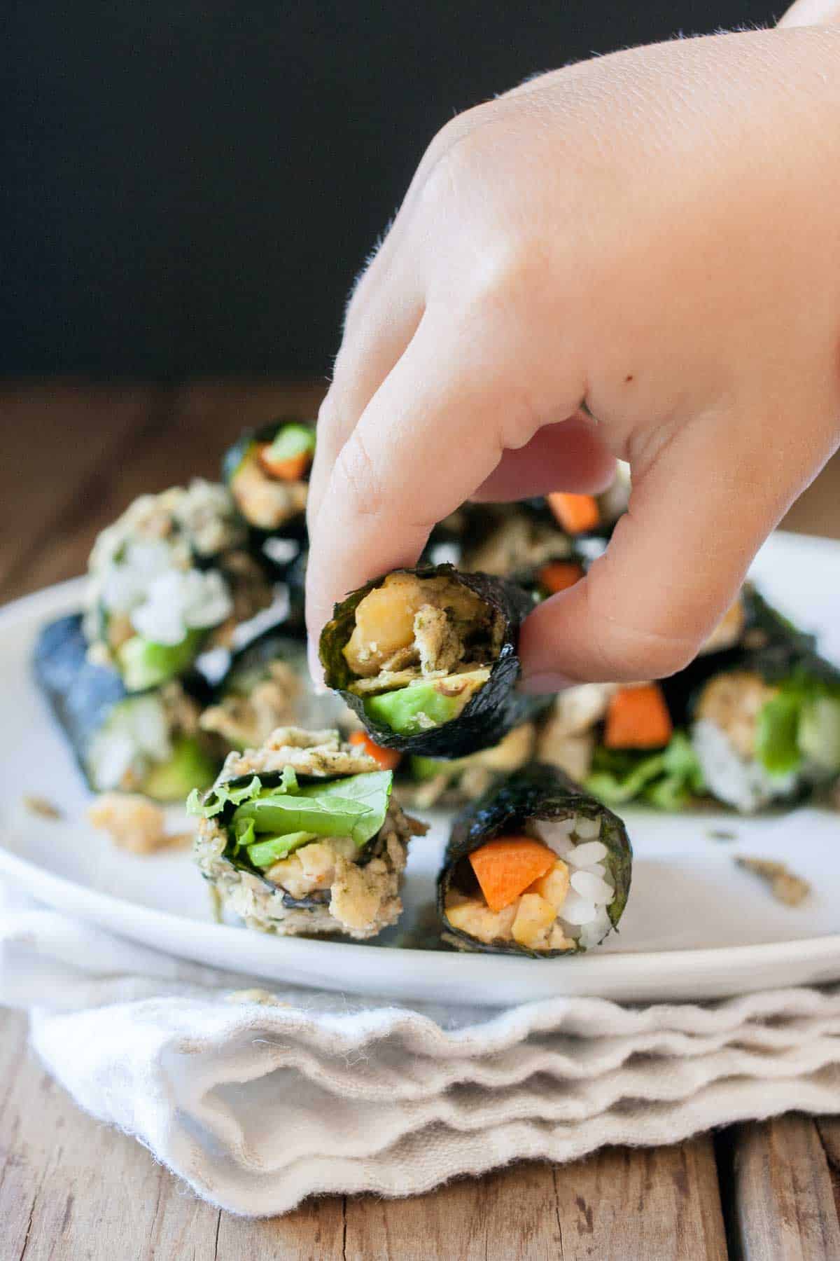 A child's hand grabbing a rolled sushi snack from a pile of them on a white plate.