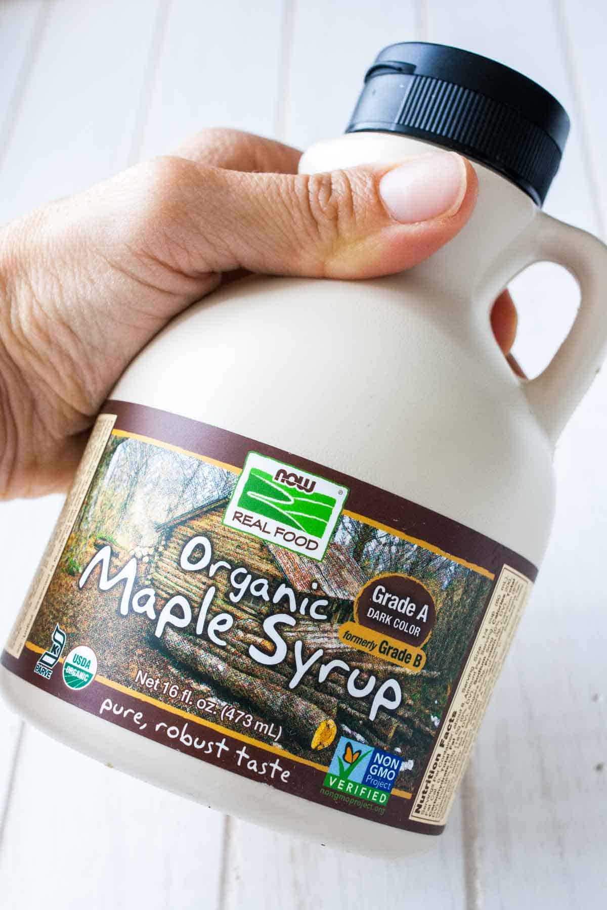 Front view of a hand holding a tan and brown bottle of maple syrup over a white wood surface.