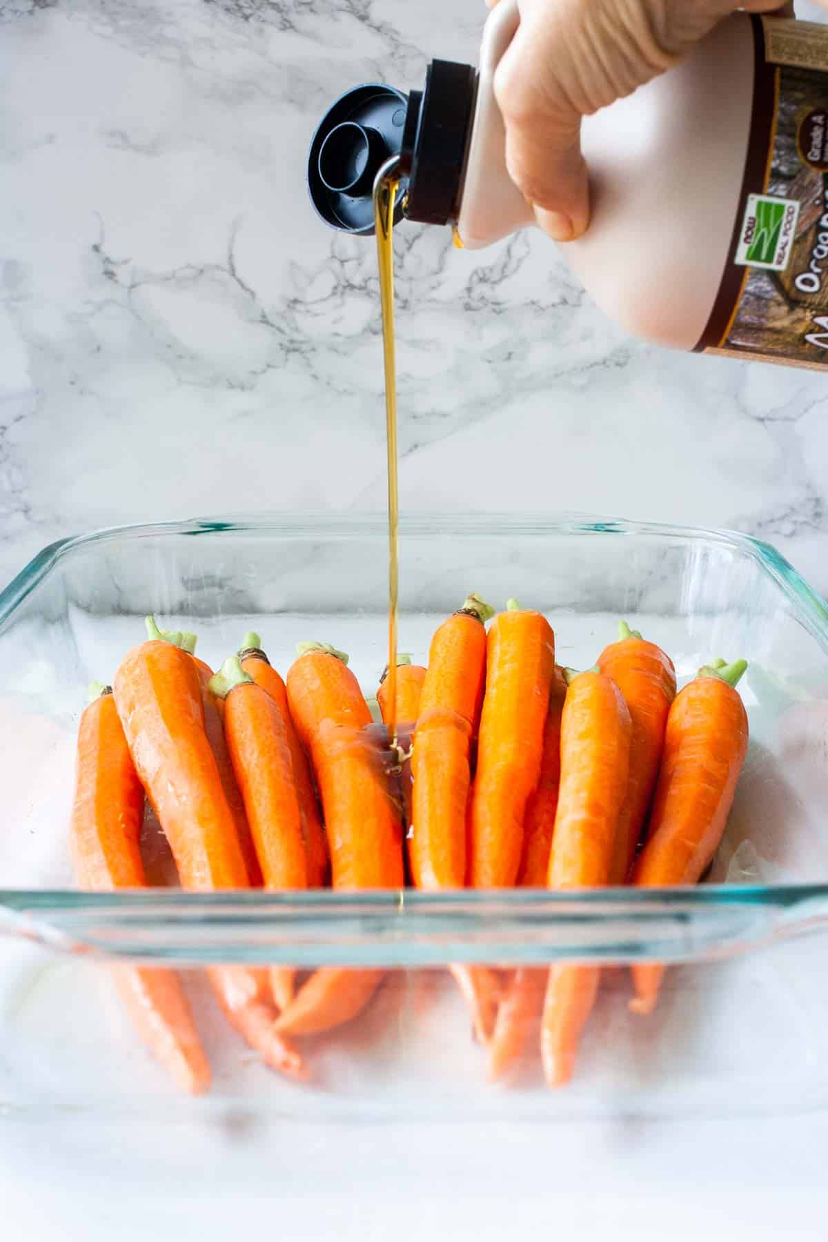 Front view of hand pouring maple syrup over carrots in a glass baking dish.