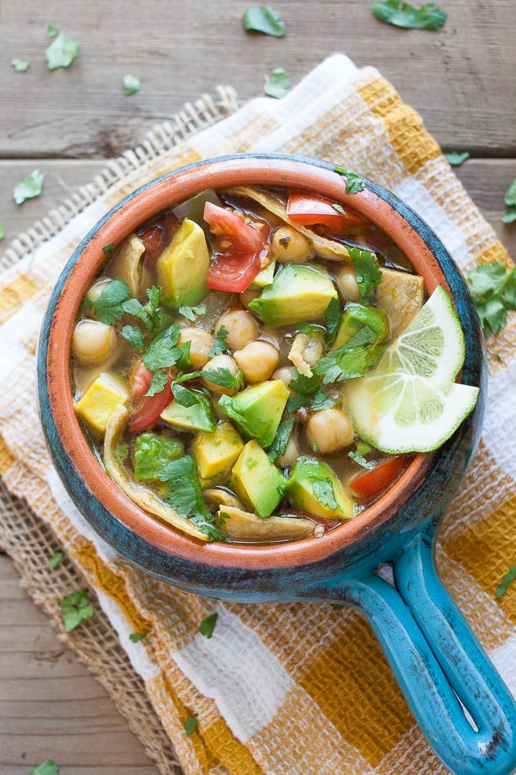 An overhead shot of vegan Chickpea Soup in a blue bowl topped with avocado and lime wedges