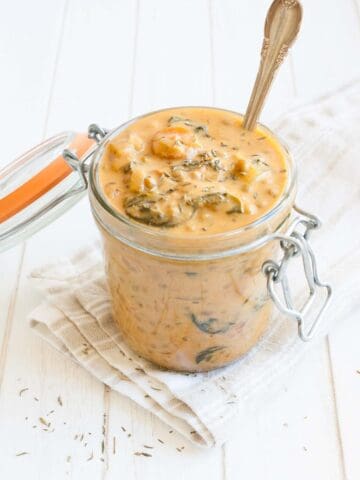 A glass jar with a pink lentil and veggie stew in it sitting on a white towel on a white wood surface.