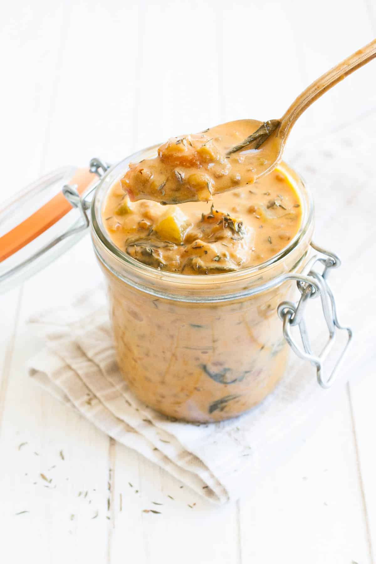 A spoon getting a bite out of a glass jar filled with creamy orange lentil veggie stew