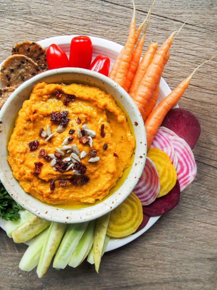 Top view of white bowl of sriracha hummus surrounded by colorful veggies and crackers