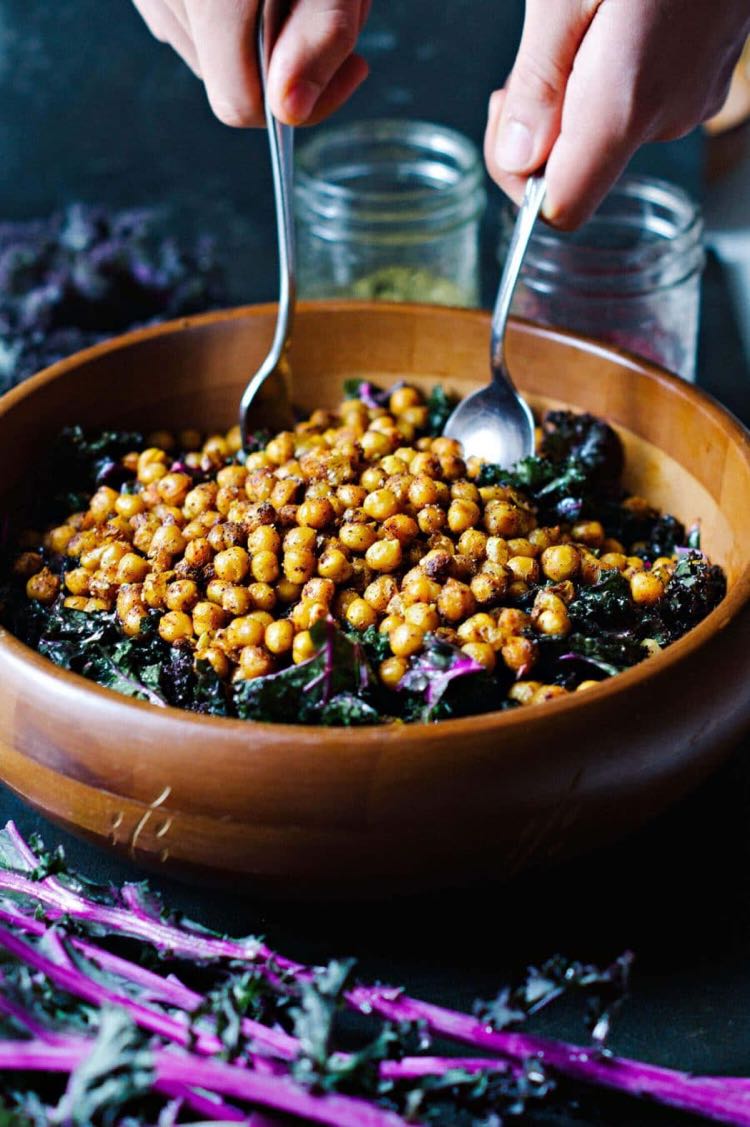 Hands holding spoons digging into brown bowl filled with kale and chickpea salad