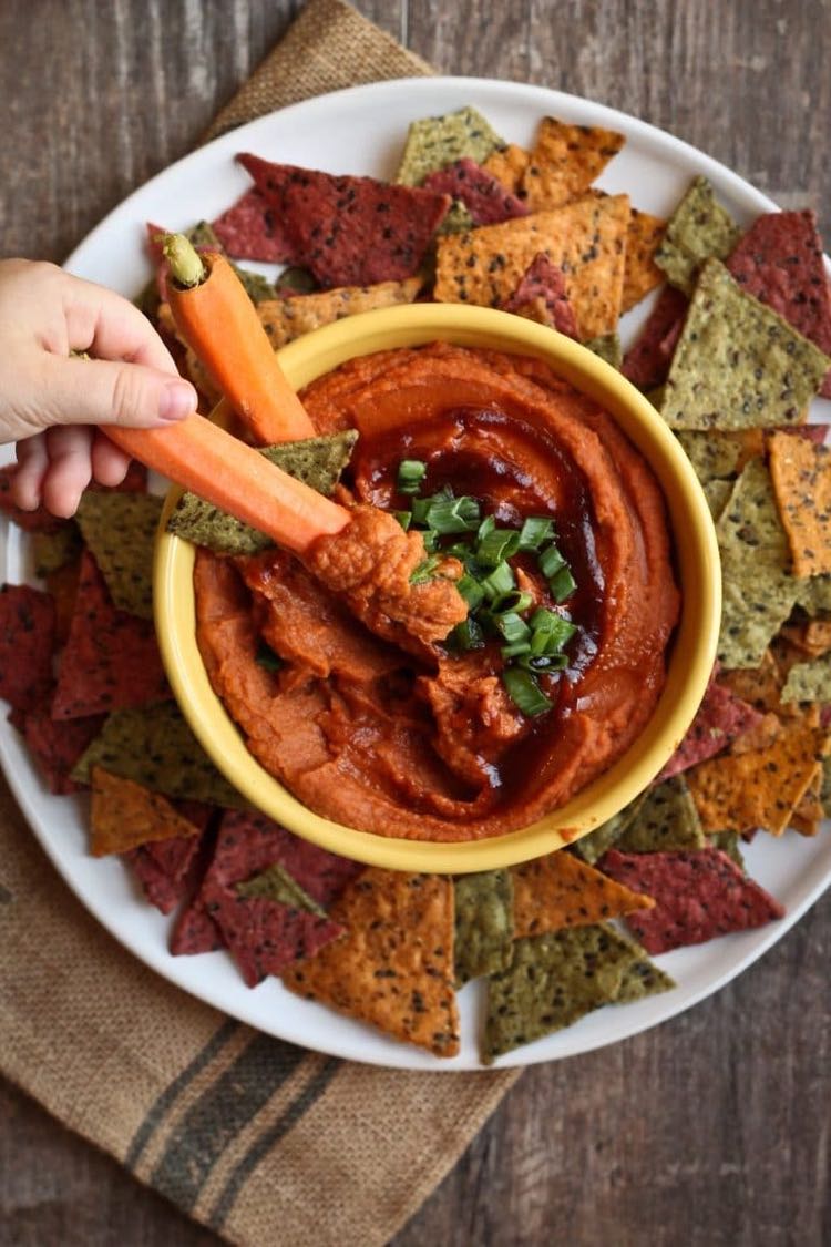 Top view of hand dipping carrot into yellow bowl of bbq hummus surrounded by chips