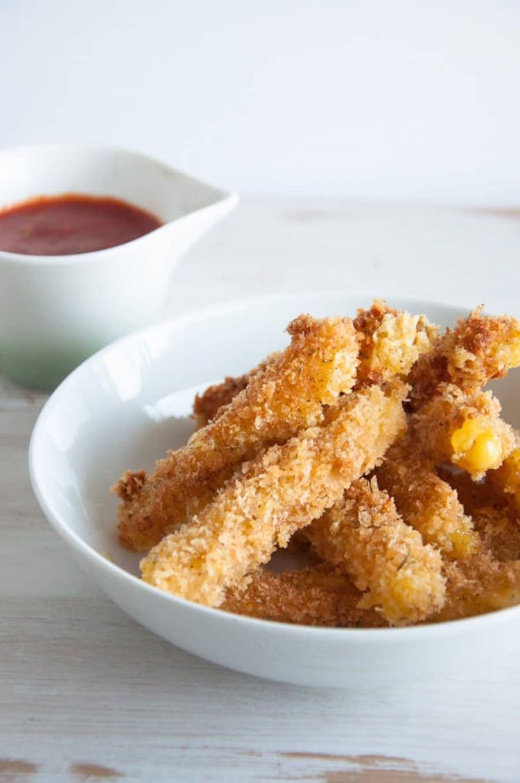 White bowl piled with crispy vegan cheese sticks in front of cup with marinara dip