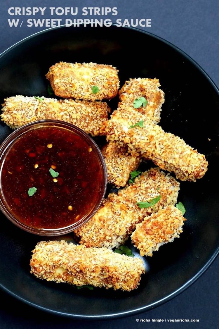 Strips of crispy covered tofu piled on a black plate with red dipping sauce