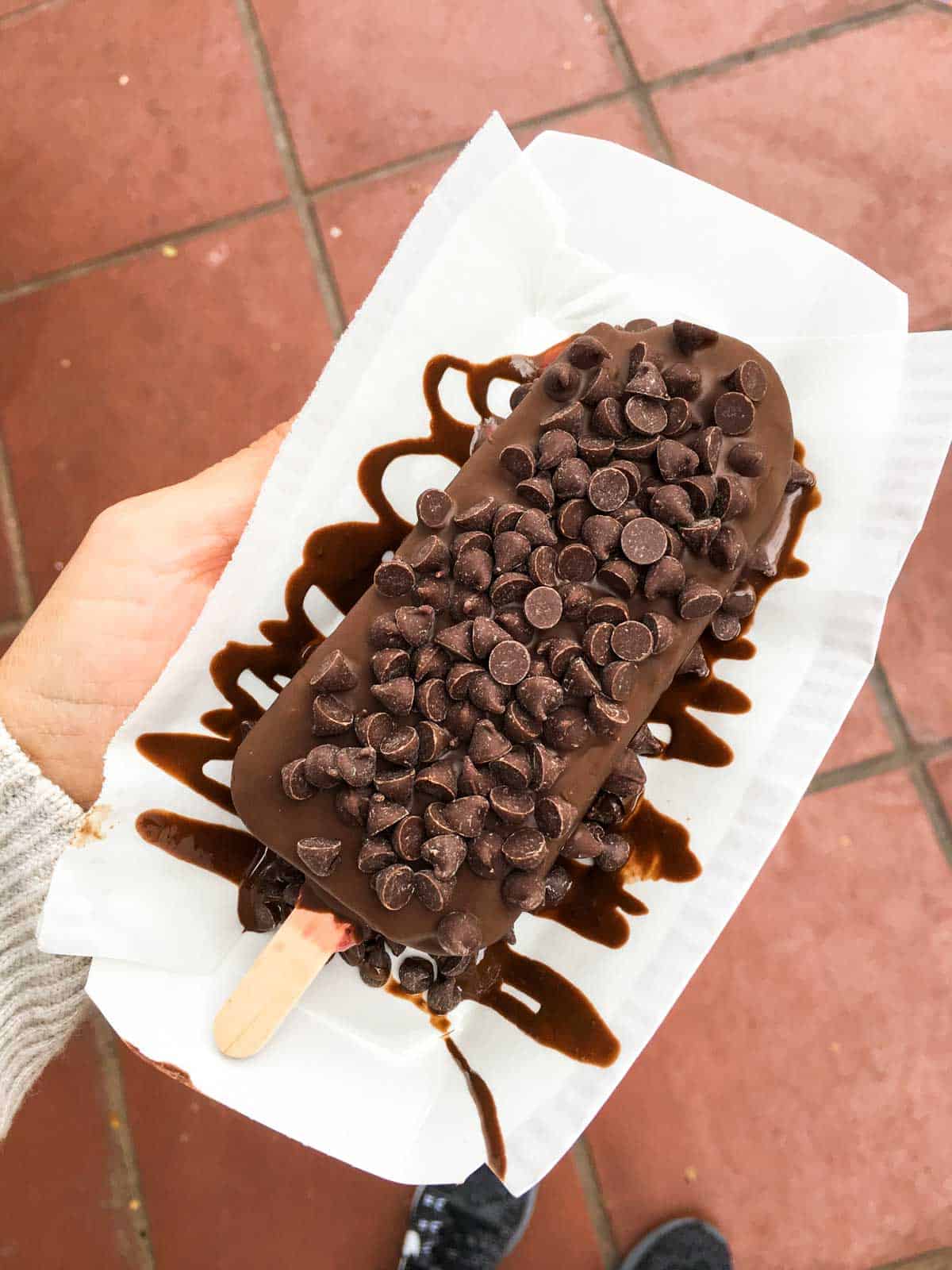 Paper container with a chocolate covered ice cream bar topped with chocolate chips.