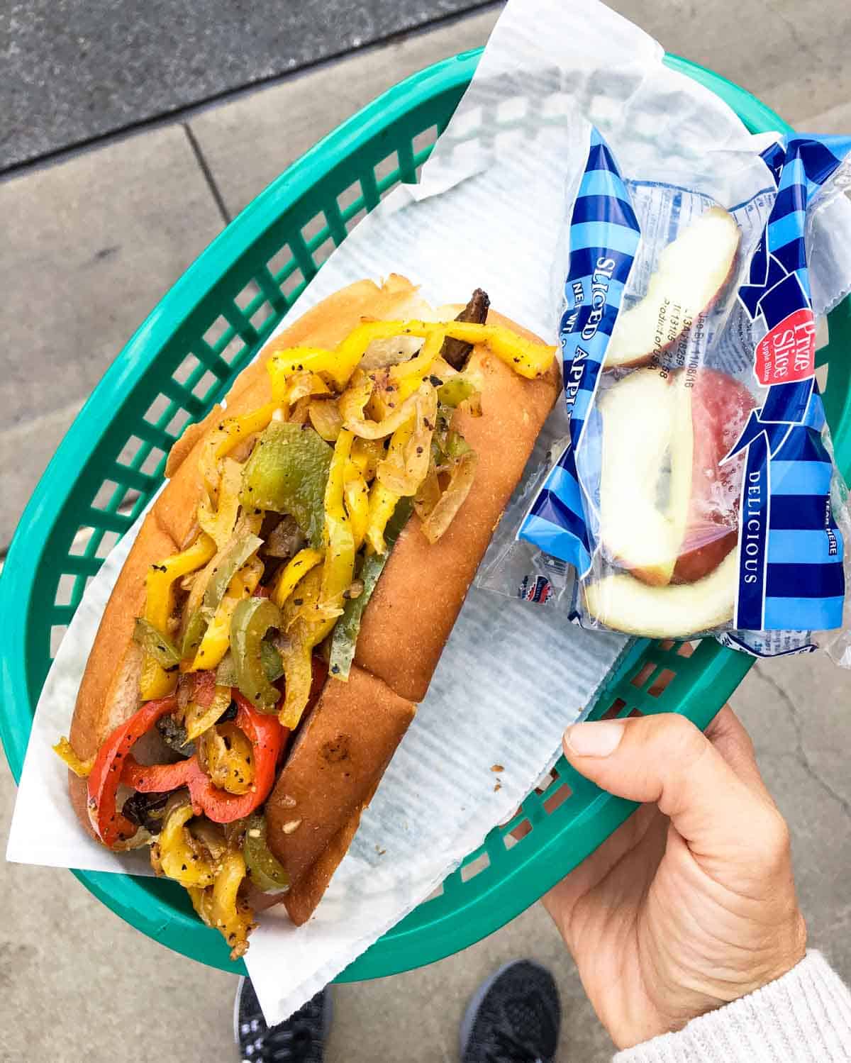 Hand holding green plastic tray with a Philly portobello sandwich and apple slices.