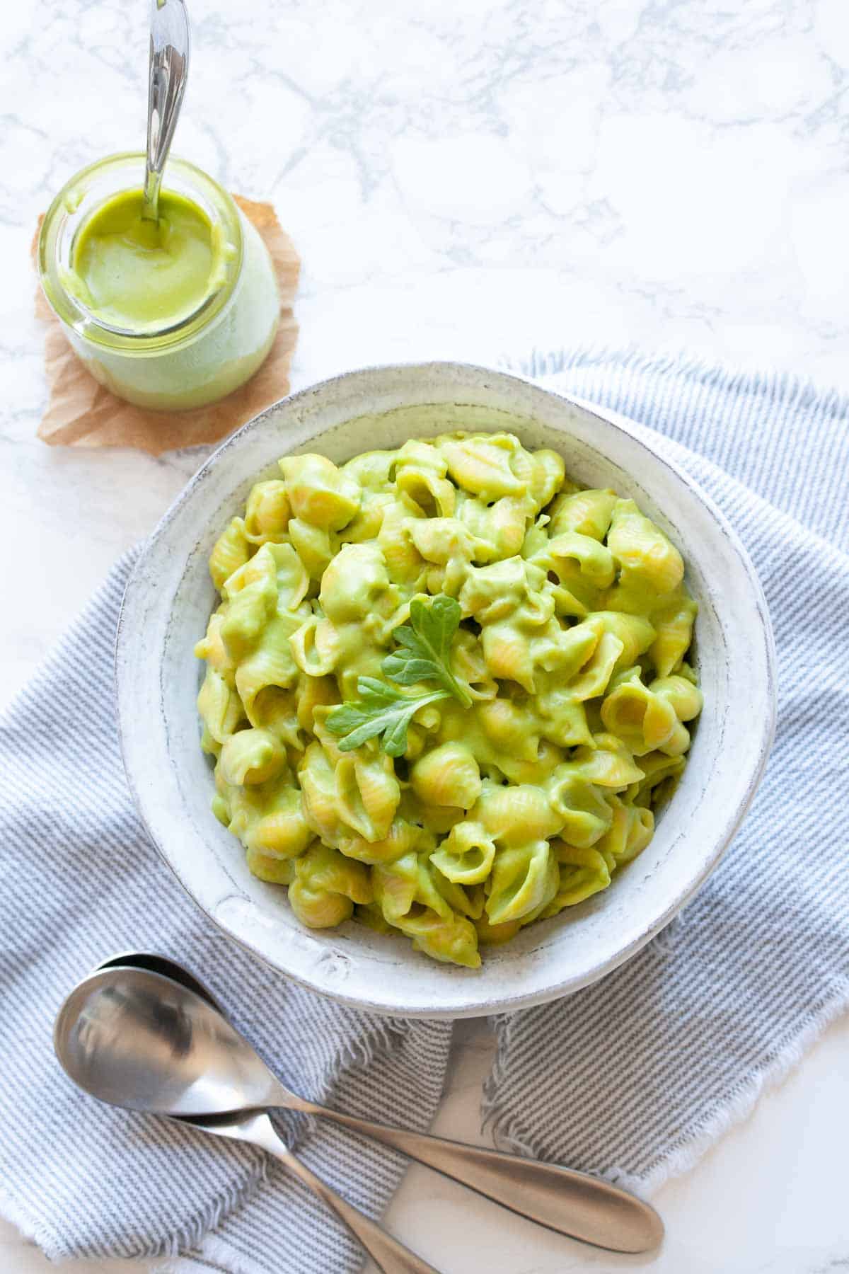 Top view of white bowl filled with green mac and cheese next to a jar of green cheese sauce..