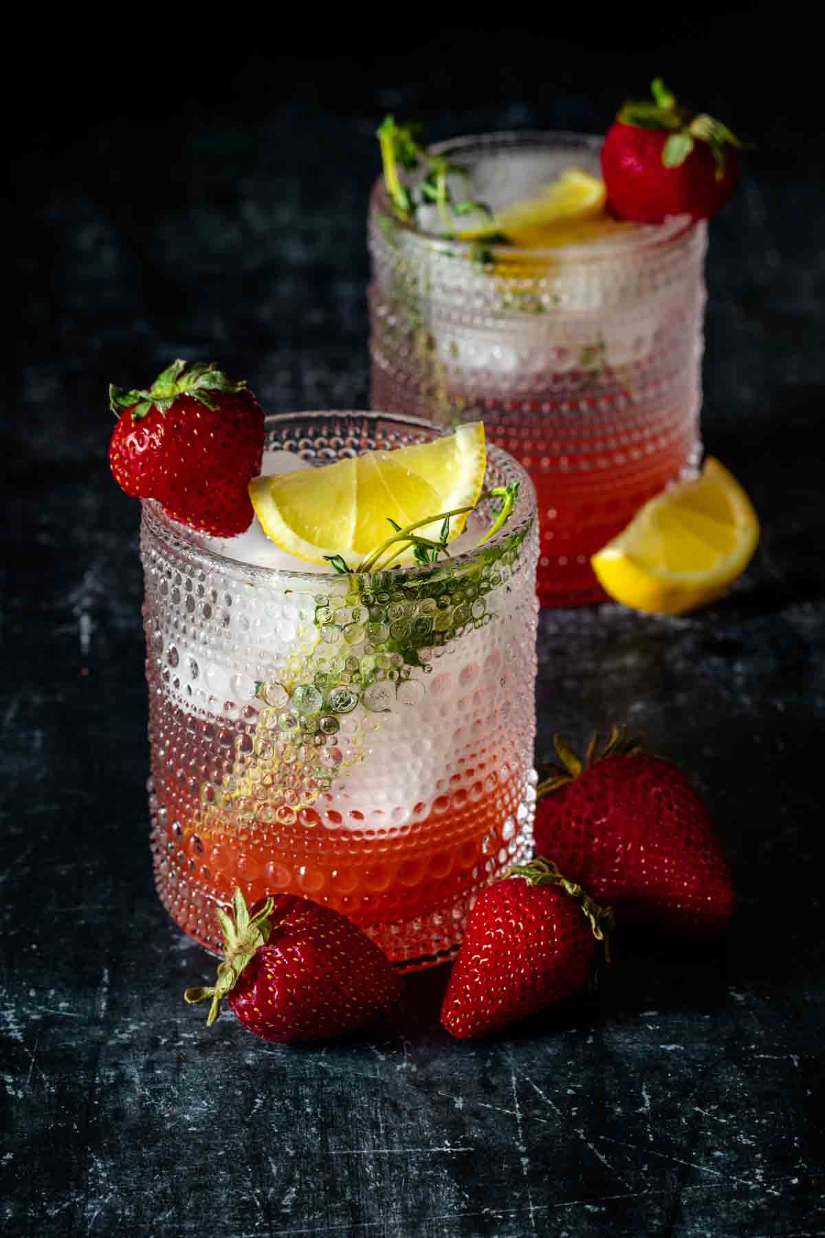 Two textured cocktail glassed with a red drink and ice topped with thyme, lemon and a strawberry.